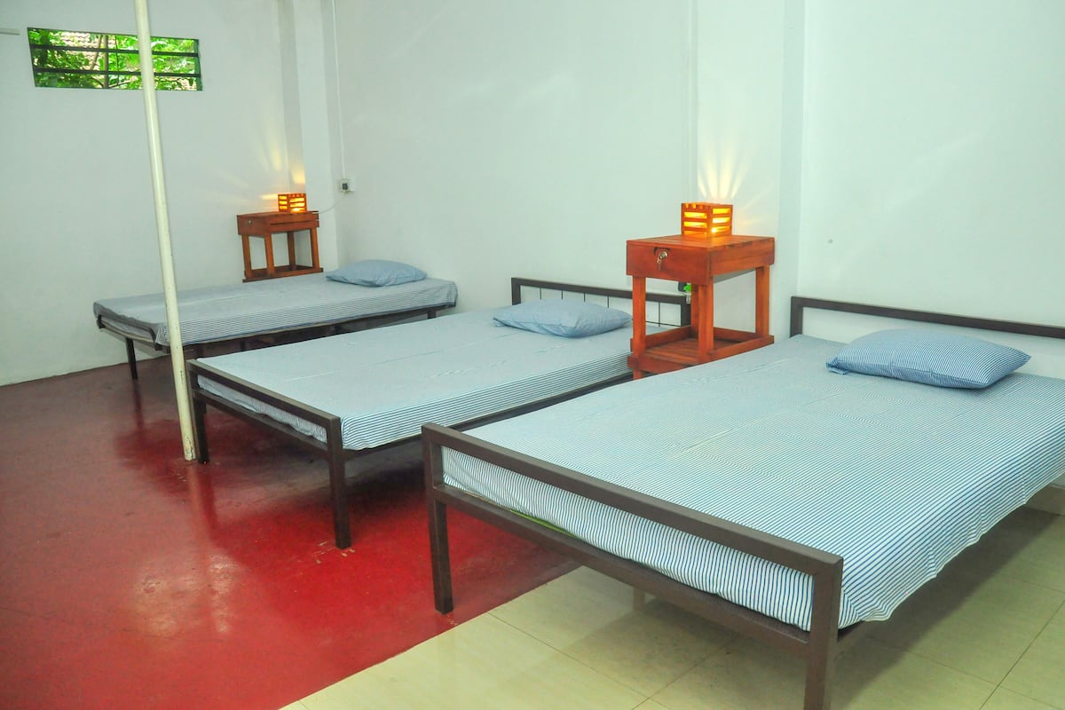 A dormitory room with three single beds, each with a wooden frame and a striped mattress. There are small wooden nightstands with lamps on each bed.