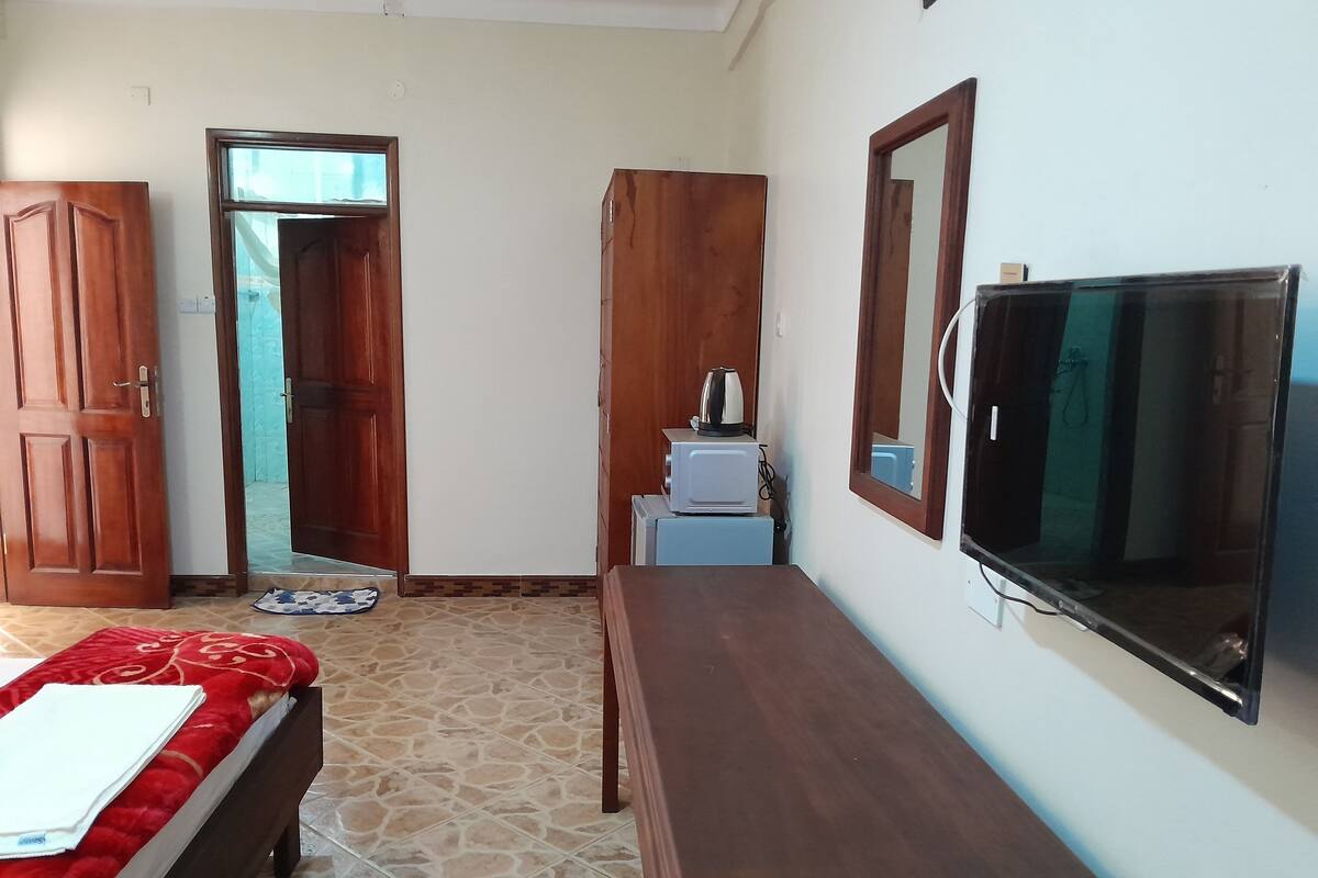 A bedroom with a bed, a wooden dresser, a television, and a small table with a kettle.