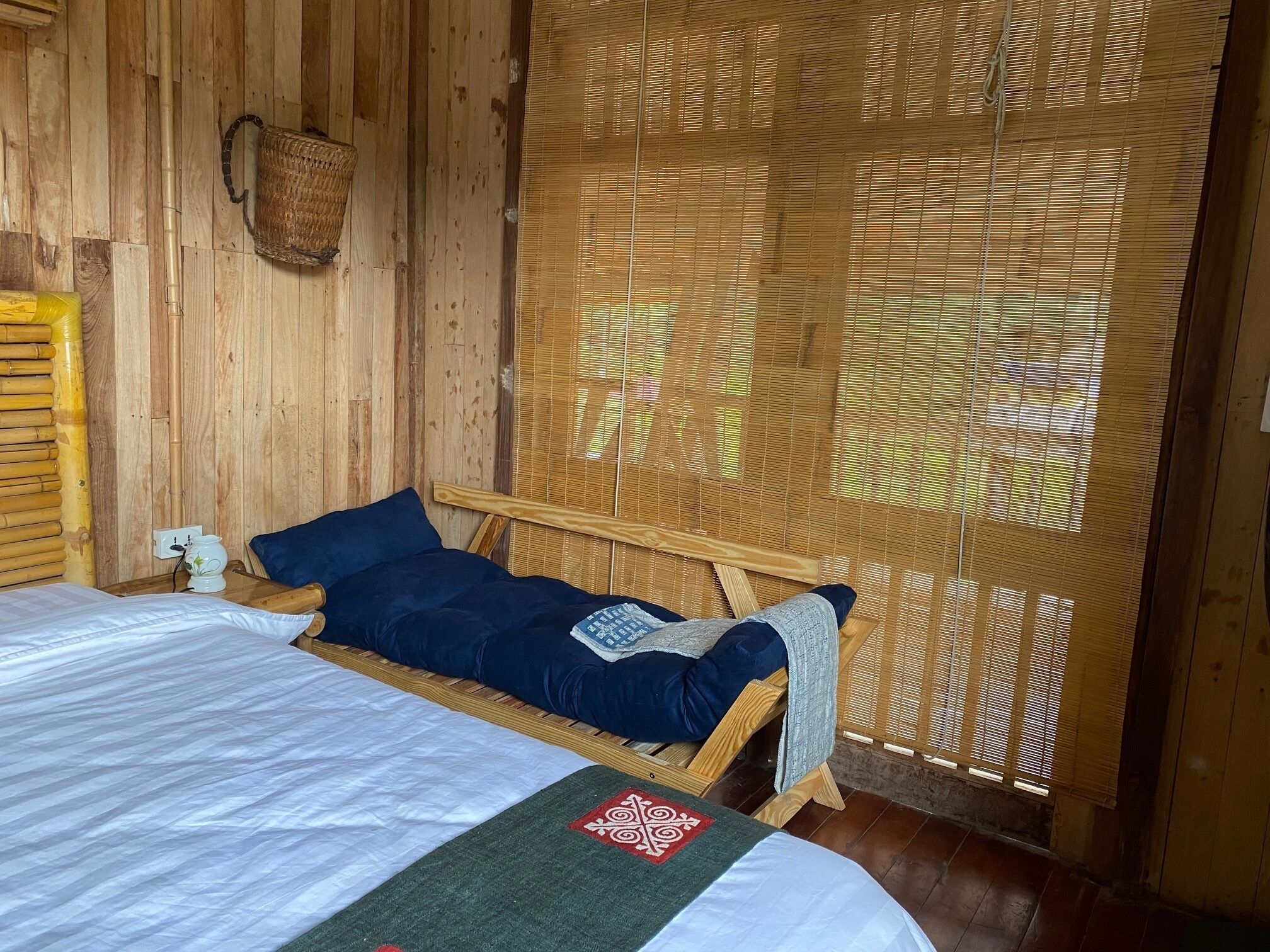Una habitación de madera con una cama, una cesta tejida y un ventanal con persianas de bambú.