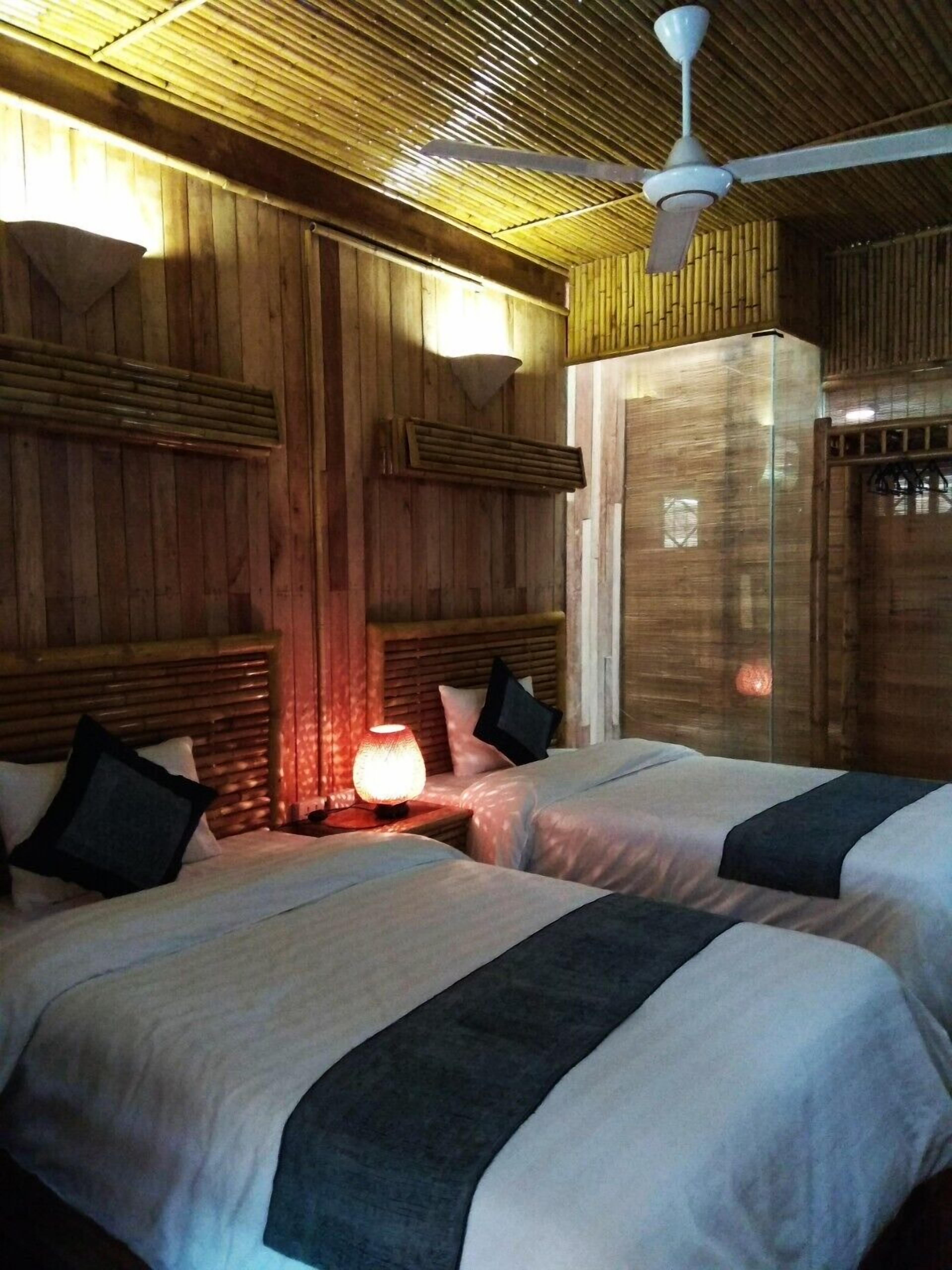 Una habitación con dos camas, paredes de madera, un ventilador de techo y una mesita de noche con lámpara.