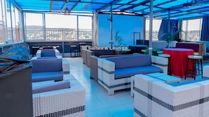 A rooftop lounge with wicker furniture, red tables, and a view of the city. - JB suites and lounge (Kampala)