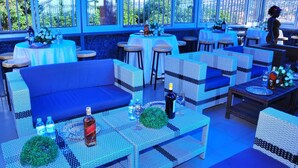 An outdoor seating area with wicker furniture, tables with drinks, and a view through large windows. - JB suites and lounge (Kampala)