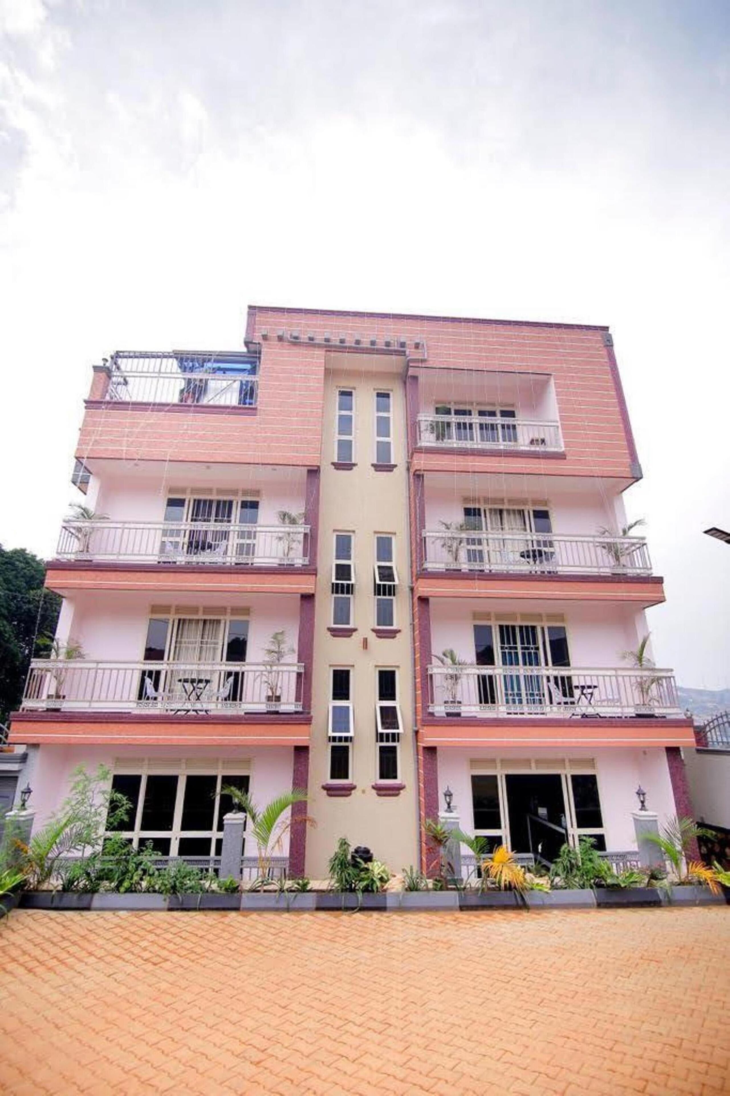 A multi-story building with balconies, a central entrance, and a paved ground area with plants.