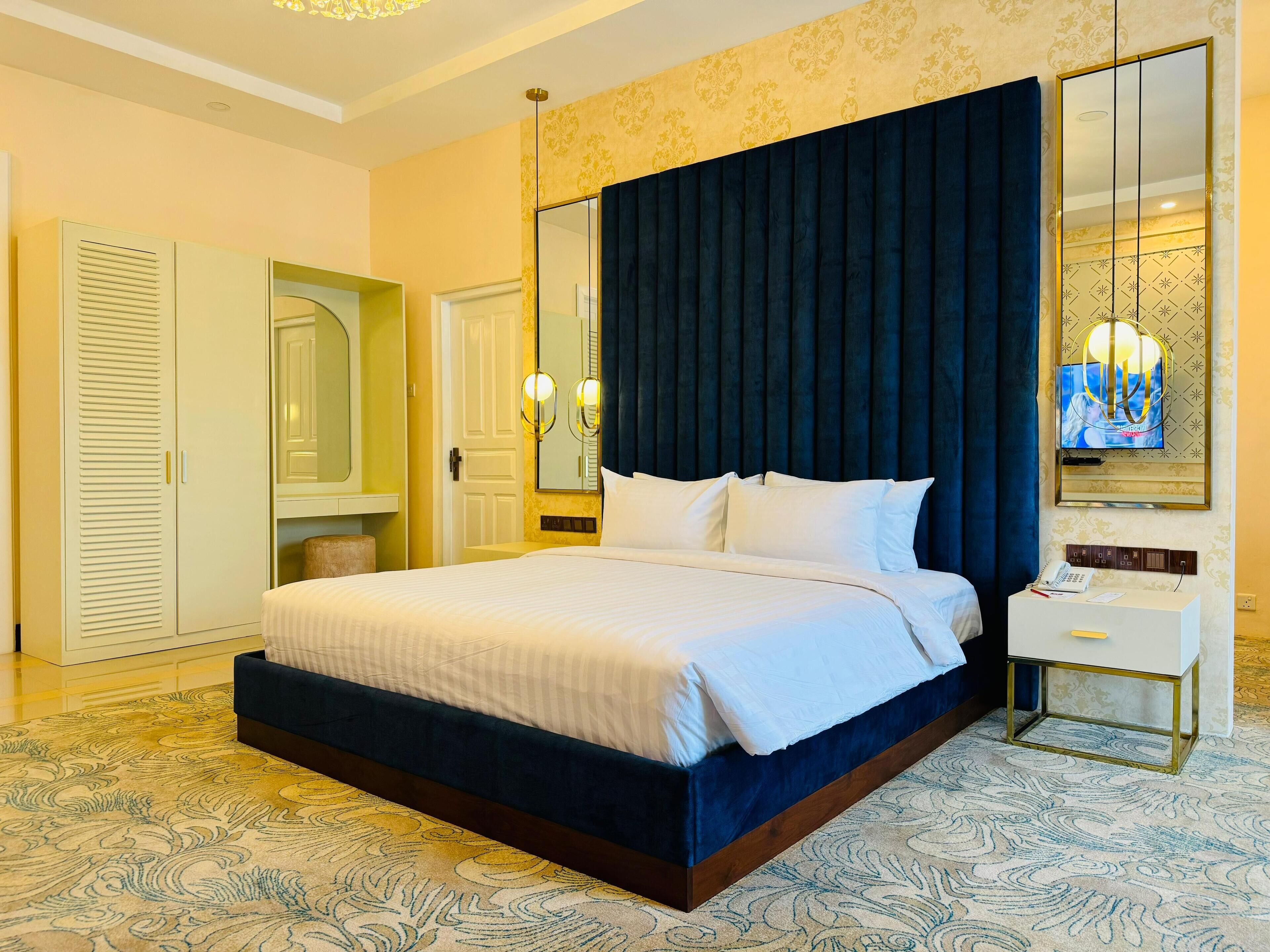 A neatly made bed with white linens, a dark blue headboard, and a large mirror above it.