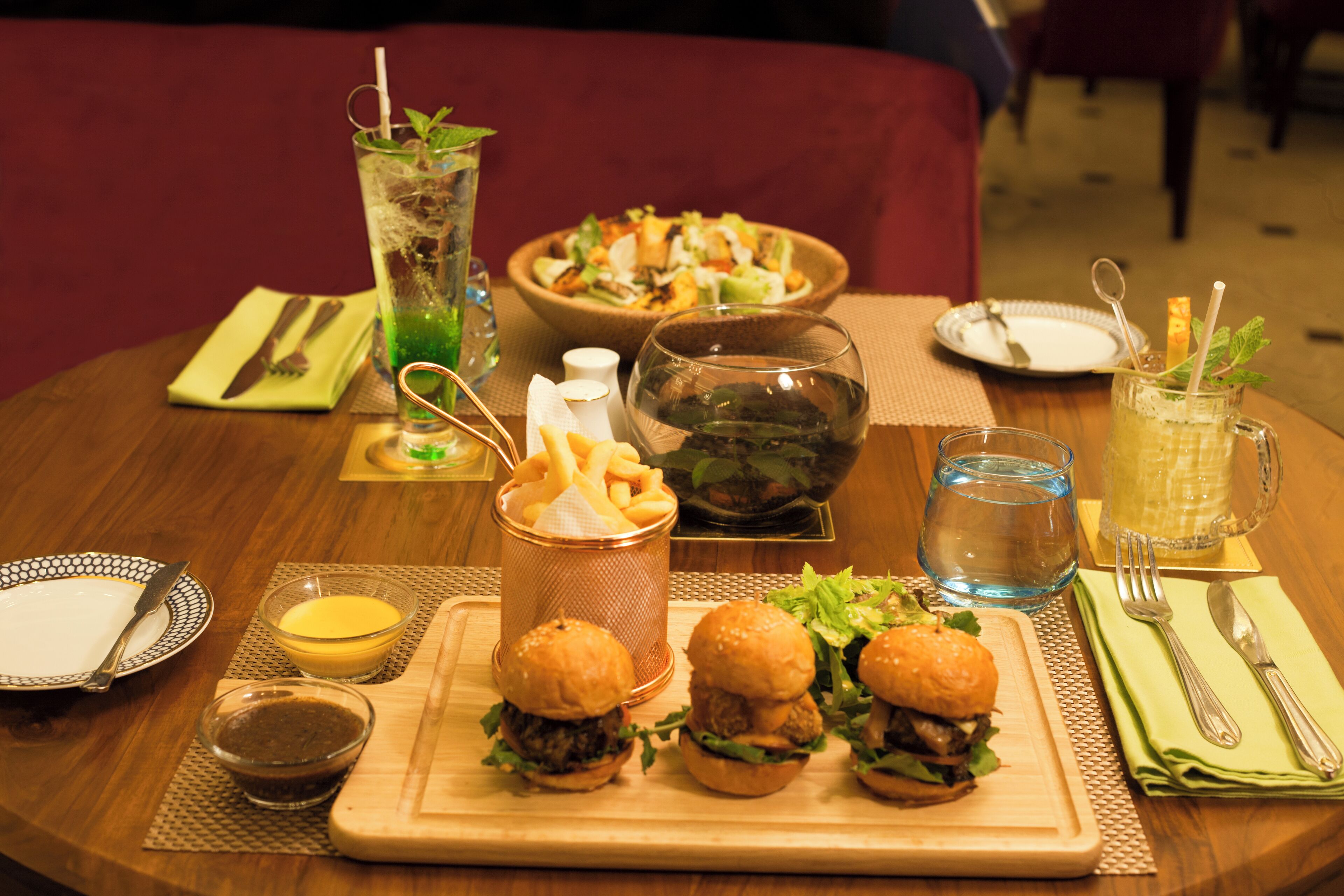 Taula de menjador amb diverses plats, com ara hamburgueses, patates fregides i una amanida, acompanyats de begudes i salses.