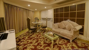 A hotel room with a red patterned carpet, a white sofa, a glass coffee table, a dining table with chairs, and a kitchen area with white cabinets and a window with blinds. - Devon Rest (Kandy)
