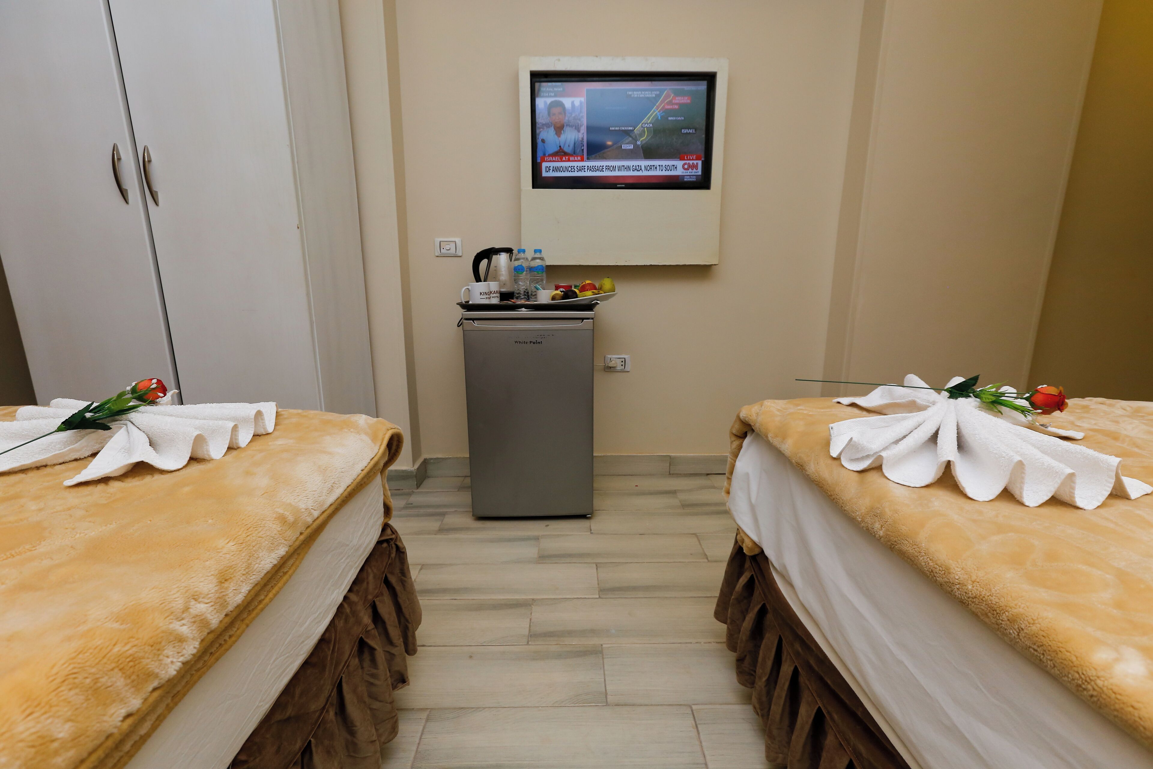 A hotel room with two beds, a small kitchenette, and a TV mounted on the wall.