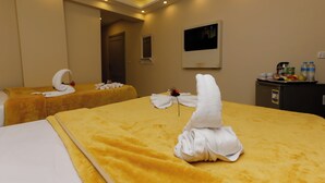 A hotel room with two beds, each with a yellow blanket and a white towel arranged in a swan shape. There is a small table with a flower and a bottle of water. - Venus Pyramids Inn (Giza)