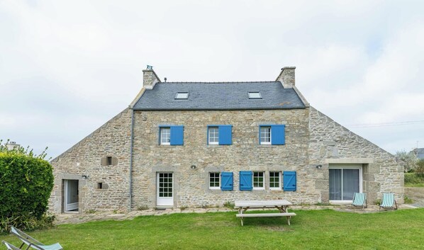 Une maison en pierre avec de nombreuses fenêtres et volets, une porte et une table de pique-nique dans le jardin.