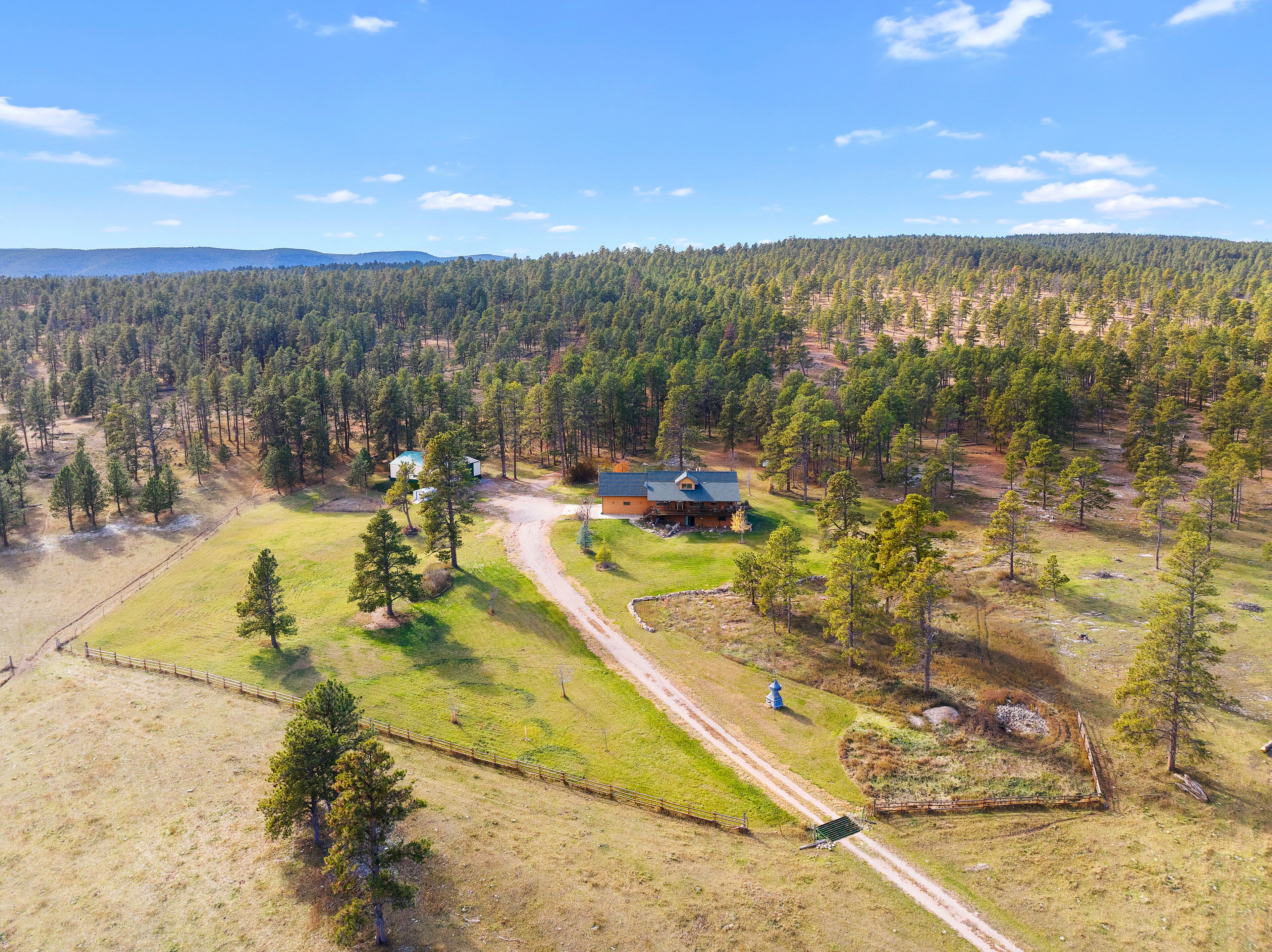 An aerial view of a rural property with a large house surrounded by dense forest, a fenced pasture, and a dirt road.