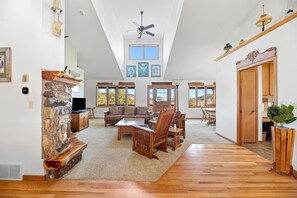 A spacious living room with a stone fireplace, wooden furniture, and large windows offering views of the outdoors. - Black Hills ranch retreat with breathtaking views, abundant wildlife, and endless room to explore, relax, and enjoy nature. (Piedmont)