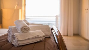 A neatly made bed with rolled white towels and a view of the sea through a window.