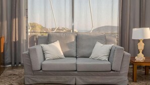A grey sofa with two cushions, a wooden side table with a lamp, and a view of a landscape through a window with curtains.
