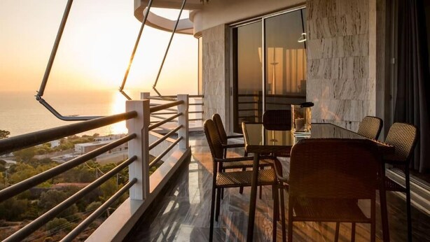 A balcony with a view of the sunset over the sea, a dining area with chairs and a table, and a modern interior with a marble wall and a glass door.