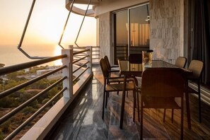 A balcony with a view of the sunset over the sea, a dining area with chairs and a table, and a modern interior with a marble wall and a glass door.