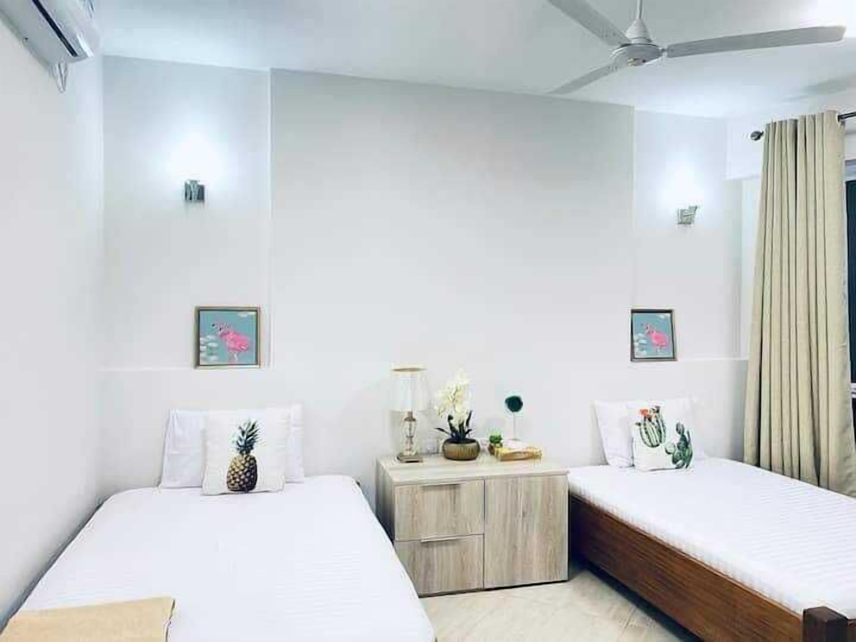 Quarto de hotel com duas camas, um ventilador de teto e uma mesa de cabeceira com luminária e objetos decorativos.