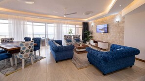 A modern living room with a dining area, featuring blue sofas, a wooden dining table, and a large TV mounted on a stone wall.