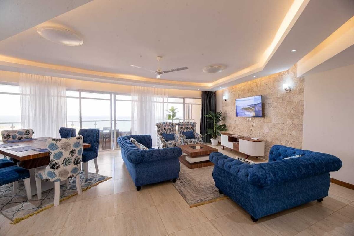 Ein modernes Wohnzimmer mit Essbereich, ausgestattet mit blauen Sofas, einem Holztisch und einem großen Fernseher an einer Steinwand.
