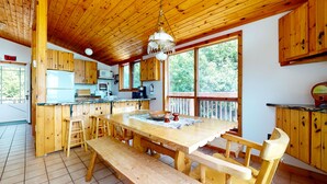 A wooden cabin kitchen with a large dining table, wooden benches, and a hanging chandelier.