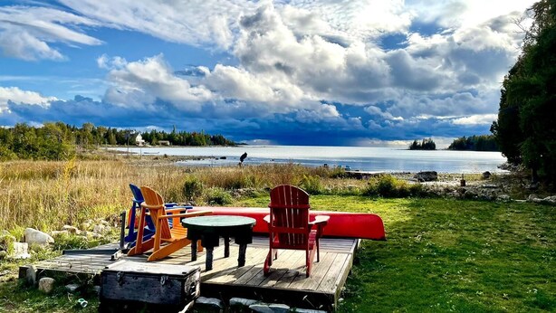 A wooden dock with chairs and a table overlooking a lake, surrounded by grassy areas and trees.