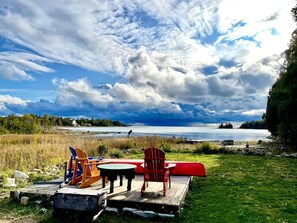 A wooden dock with chairs and a table overlooking a lake, surrounded by grassy areas and trees.