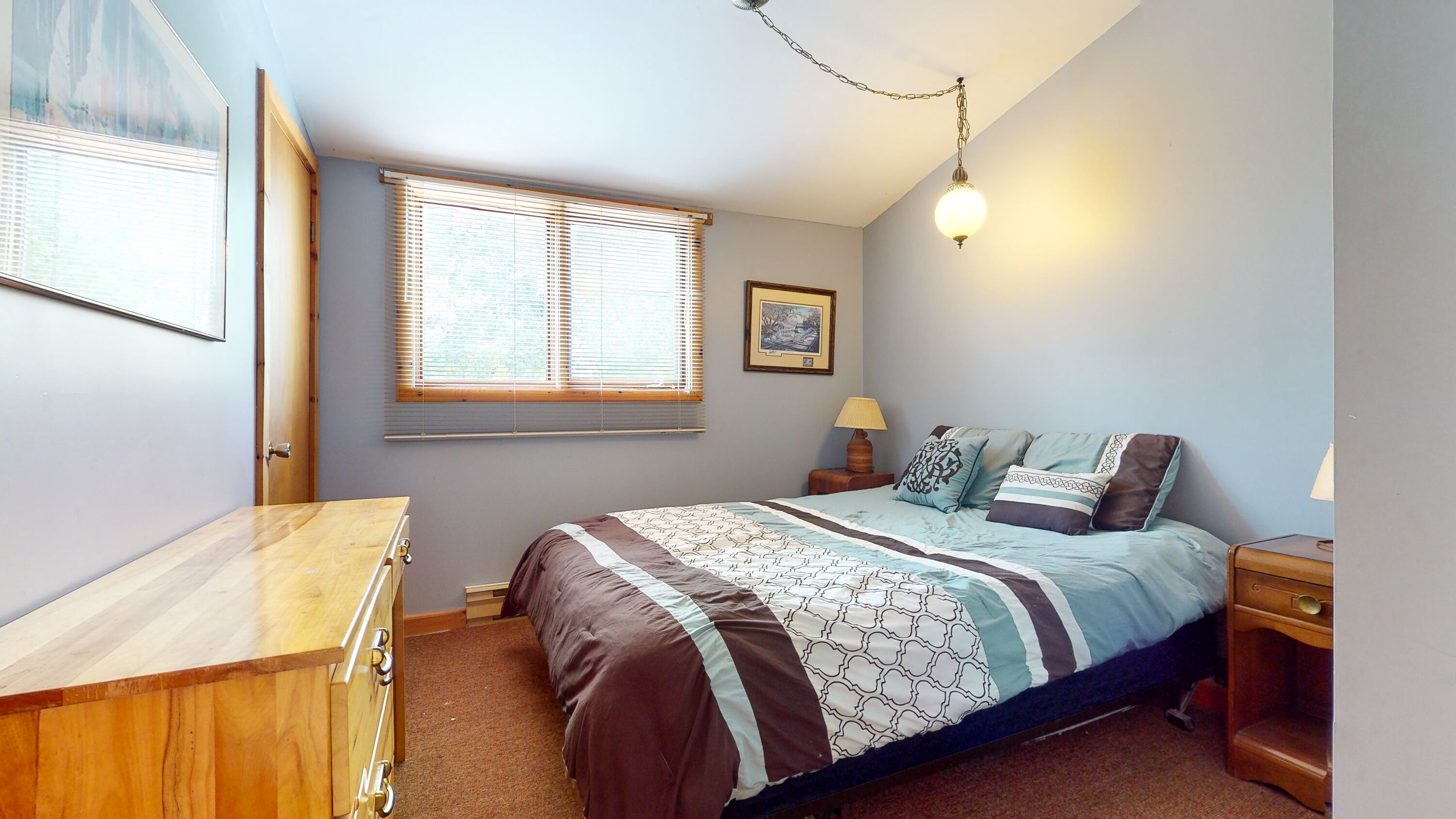 Une chambre à coucher comprenant un lit, une commode en bois, une table de chevet, une lampe, une fenêtre avec des stores et un luminaire au plafond.