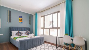 A bedroom with a bed, a desk, two chairs, a window with curtains, and wall art.