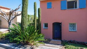 Un bâtiment rose avec une porte noire, deux fenêtres dotées de volets bleus et une petite fenêtre avec un grillage métallique.