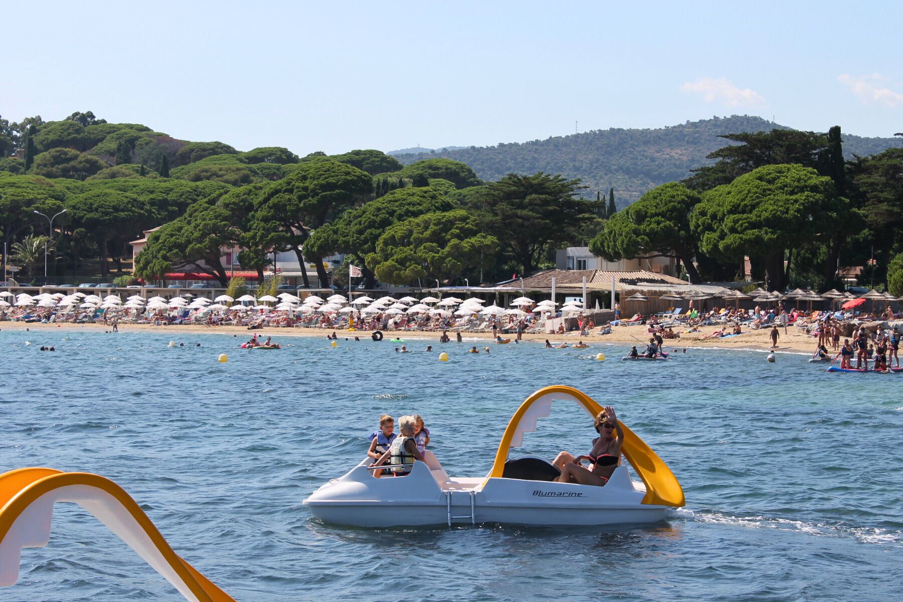 Un bote con dos pasajeros, una playa con sombrillas y gente, y un fondo de árboles y colinas.