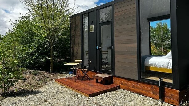 A modern cabin with a large window, a wooden deck, and a bed inside.
