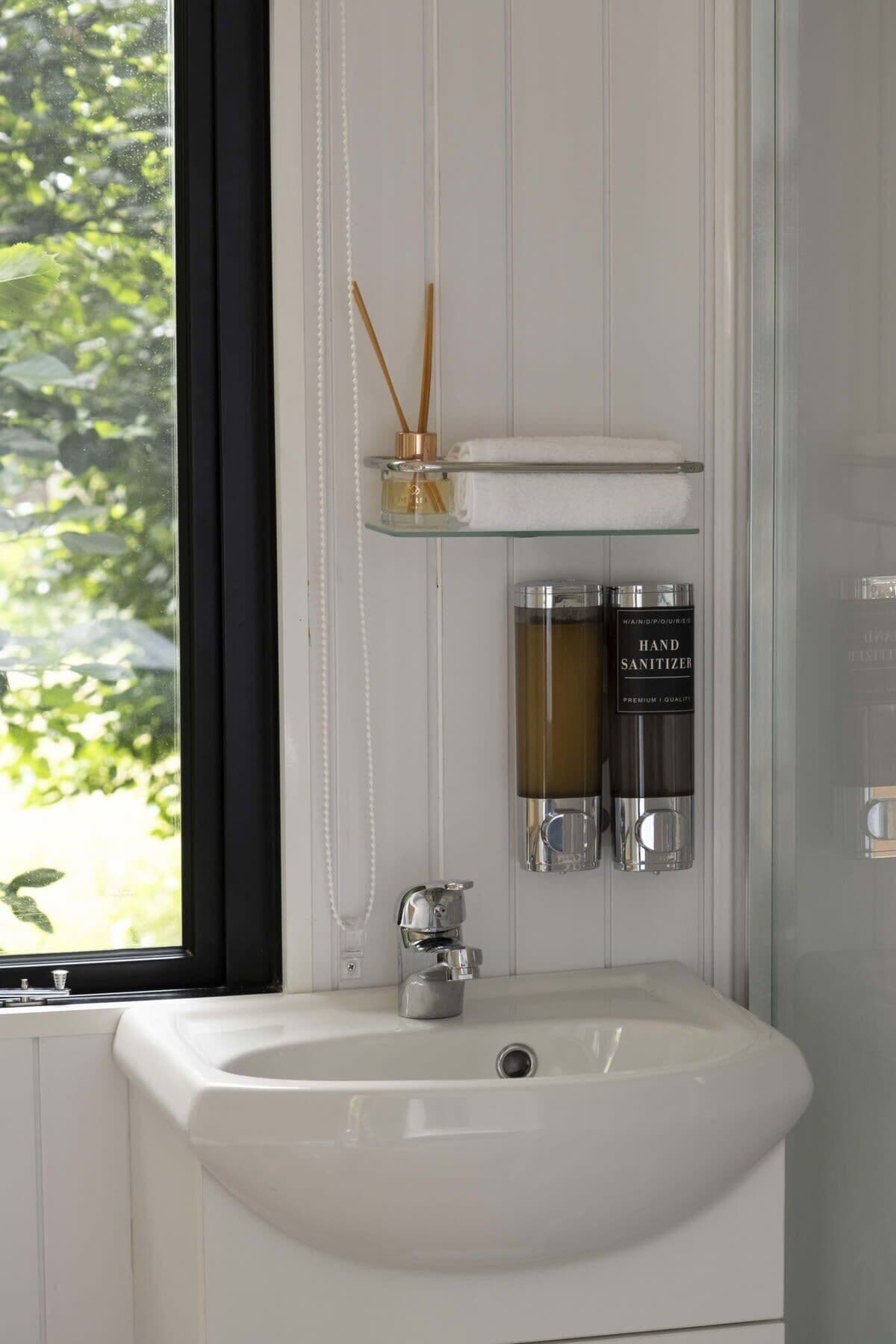 A bathroom with a white sink, a glass shelf with a bottle and a towel, and a hand sanitizer dispenser.