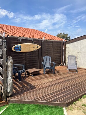A wooden deck with a surfboard, two chairs, and a table. - Villa du Raz 2, Traditionnelle et Familiale à 40 Mètre de la Piste Cyclable (Mimizan)