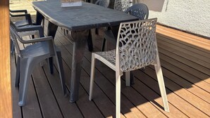 A covered outdoor patio with a table and chairs, a grill, and a view of a building in the background. - Villa du Raz 2, Traditionnelle et Familiale à 40 Mètre de la Piste Cyclable (Mimizan)