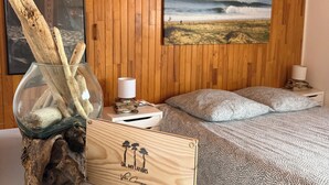 A wooden-paneled room with a bed, a framed picture of a beach, and a glass bowl with driftwood. - Villa du Raz 2, Traditionnelle et Familiale à 40 Mètre de la Piste Cyclable (Mimizan)