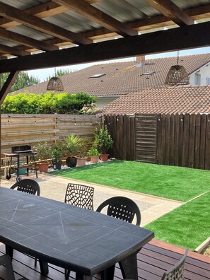 A patio with a table and chairs, a wooden deck, a fenced backyard with artificial grass, and a tiled roof. - Villa du Raz 2, Traditionnelle et Familiale à 40 Mètre de la Piste Cyclable (Mimizan)