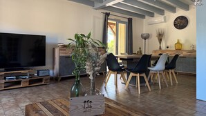 A living room with a TV, wooden coffee table, and a dining area with chairs and a table. - Villa du Raz 2, Traditionnelle et Familiale à 40 Mètre de la Piste Cyclable (Mimizan)