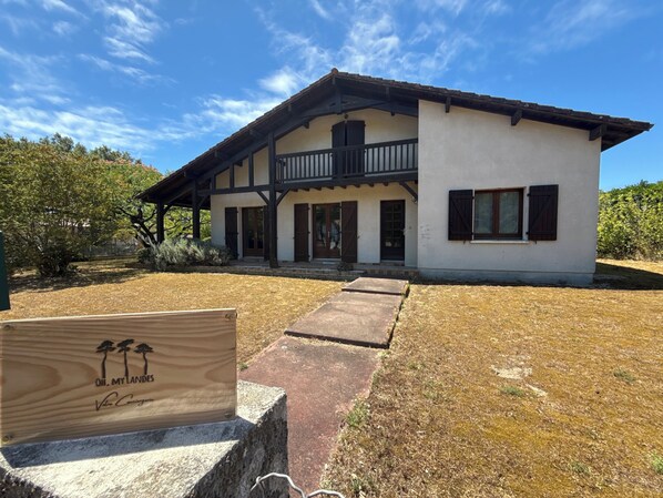 A two-story building with a balcony, a wooden sign in front, and a dry lawn. - Villa du Raz 2, Traditionnelle et Familiale à 40 Mètre de la Piste Cyclable (Mimizan)