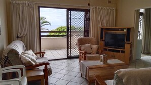 A living room with a TV, wooden furniture, a balcony with curtains, and a view of the sea. - 39B Club Mykonos (eMdloti)