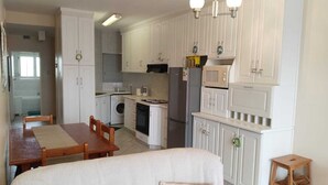 A compact kitchen with white cabinetry, a built-in oven, microwave, and a refrigerator. There is a dining area with a wooden table and chairs. - 39B Club Mykonos (eMdloti)