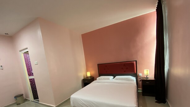 Una habitación de hotel con una cama grande, dos mesitas de noche con lámparas, una puerta con cortina y un cubo de basura.