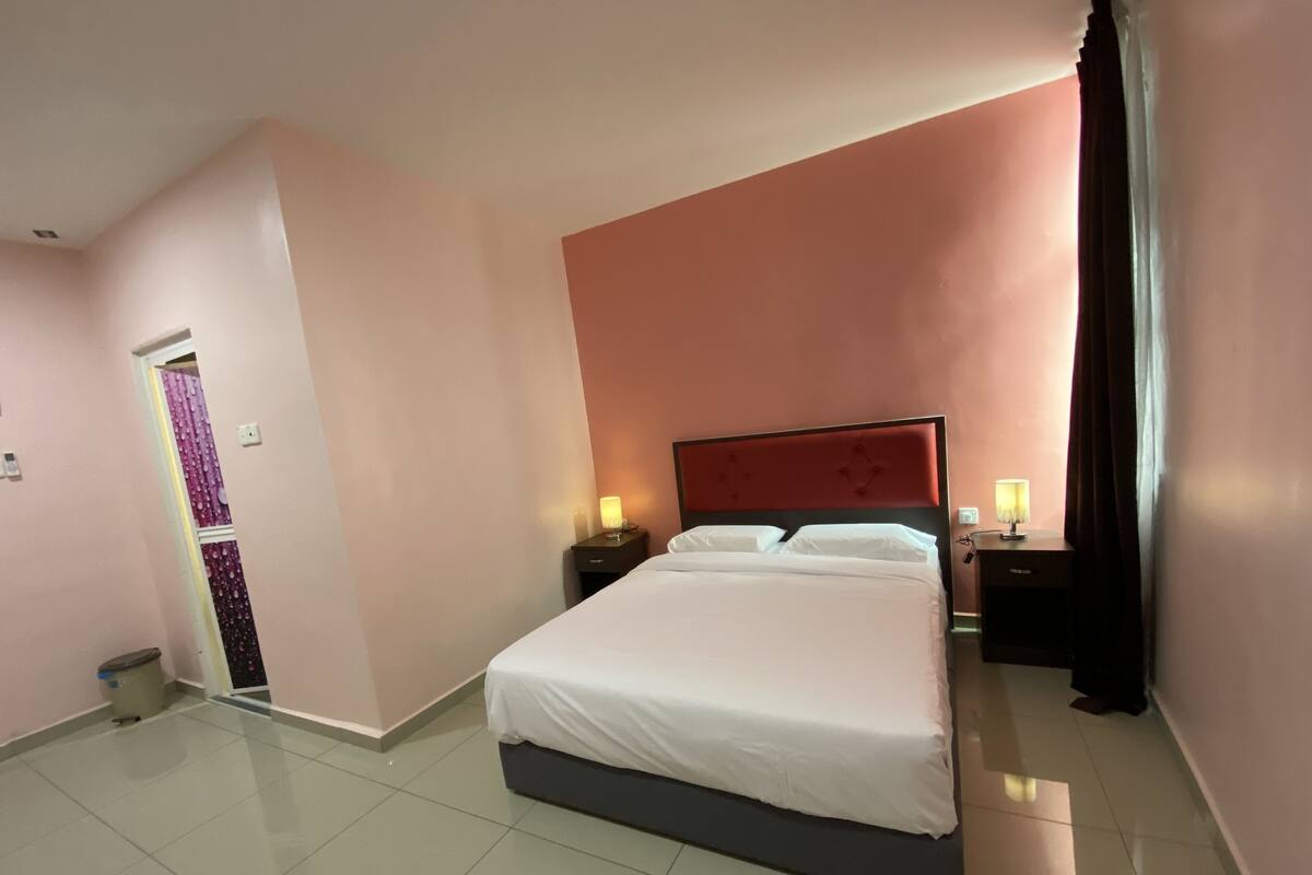 Una habitación de hotel con una cama grande, dos mesitas de noche con lámparas, una puerta con cortina y un cubo de basura.