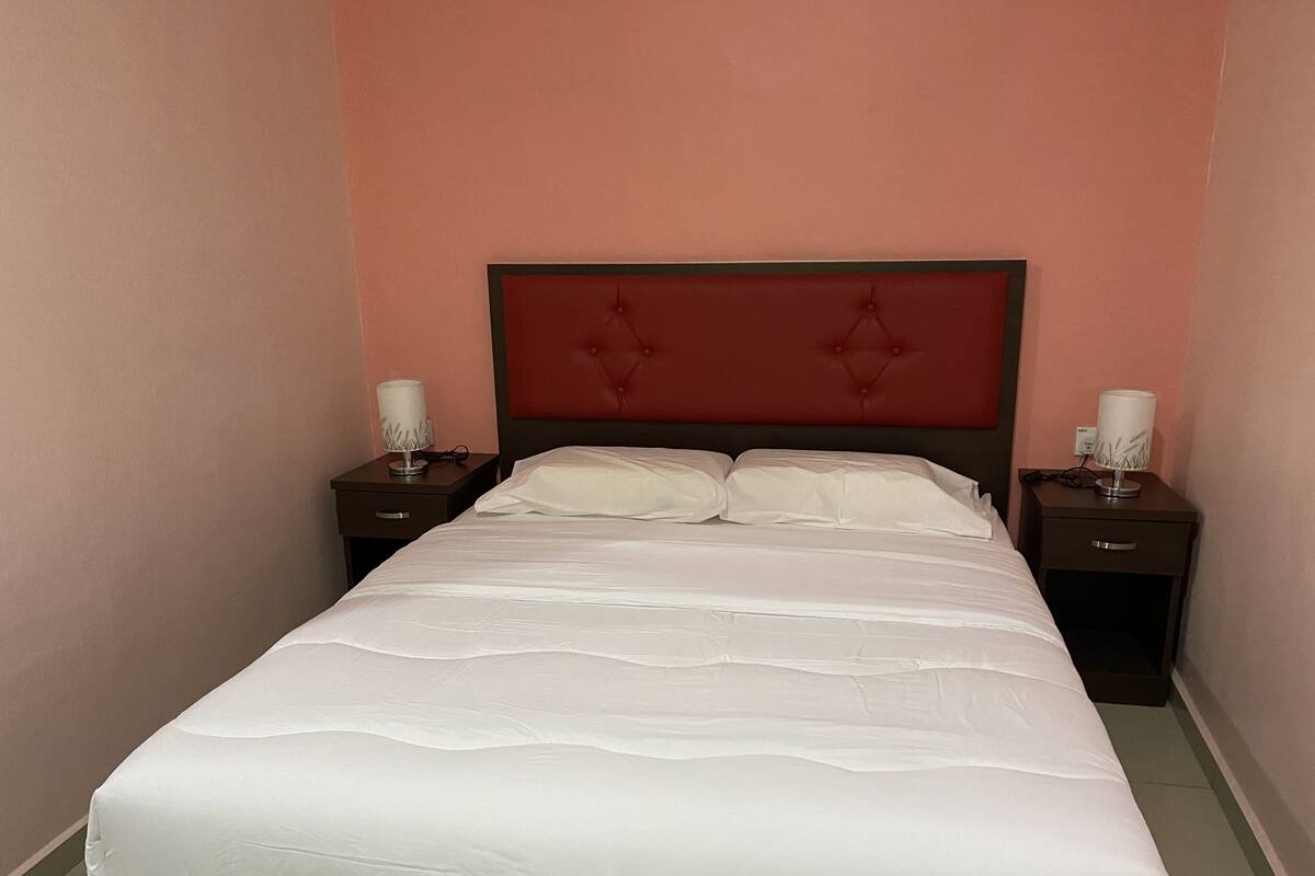 Una cama bien hecha con ropa de cama blanca en una habitación con paredes rosas y un cabecero rojo. Hay dos mesitas de noche con lámparas a cada lado de la cama.