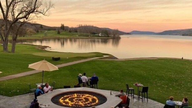 An outdoor seating area with a fire pit surrounded by tables and chairs, overlooking a lake. - Score The perfect Summer Winter getaway: Lakeside retreat for your winning team  (Maryland)