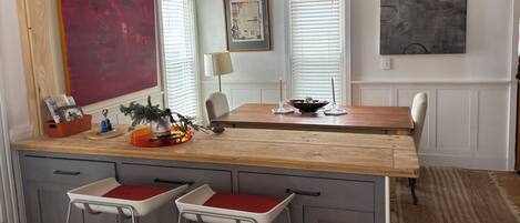 A kitchen with a wooden countertop, two bar stools, a dining table, and a large painting on the wall.