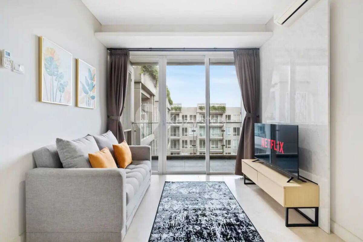 Ein modernes Wohnzimmer mit grauem Sofa, einem schwarz-weißen Teppich, einem Fernseher auf einem Holzboden und einem Balkon mit Blick auf die Stadt.
