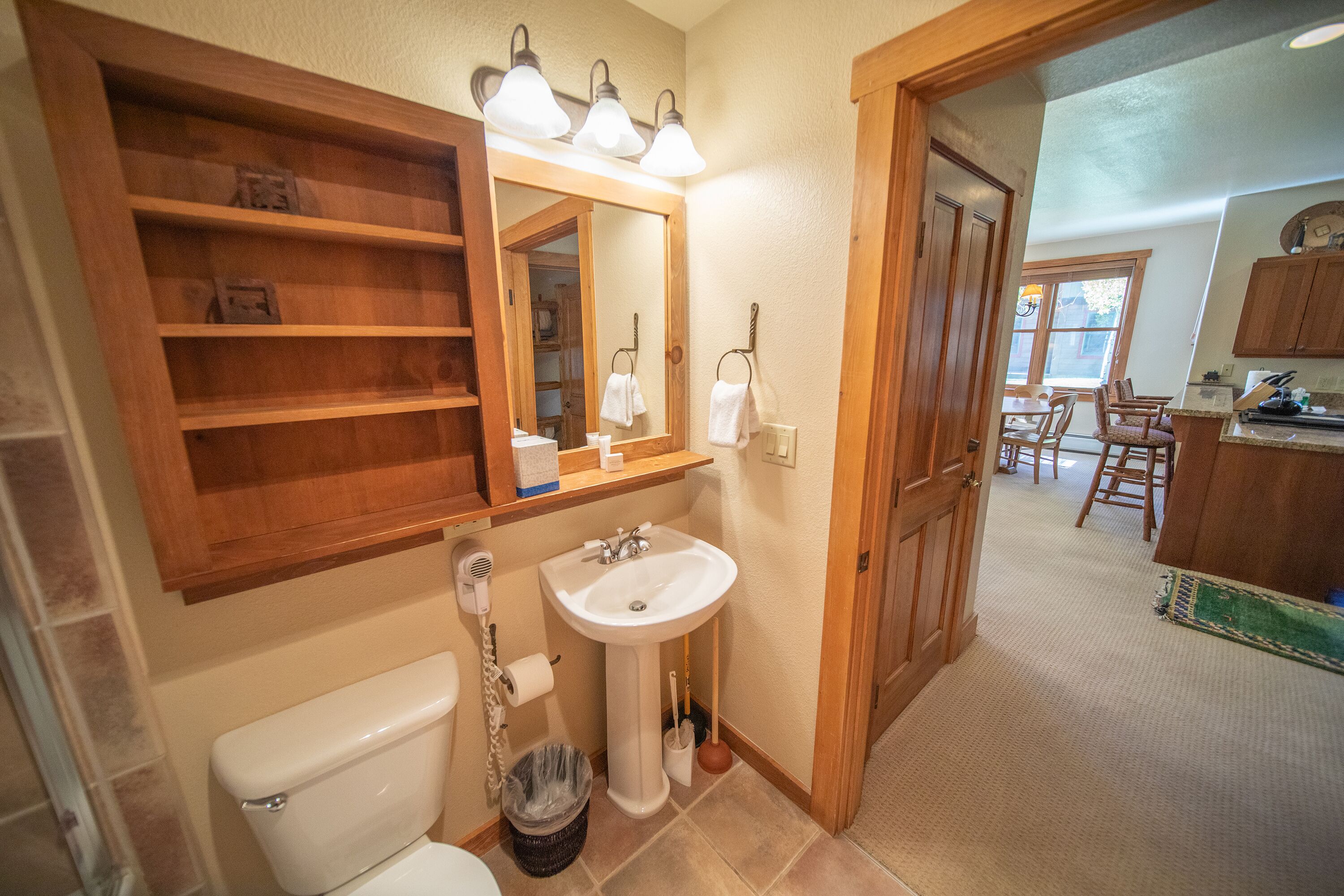 A bathroom with a toilet, sink, and mirror. There is a wooden shelf above the sink and a mirror above the toilet.
