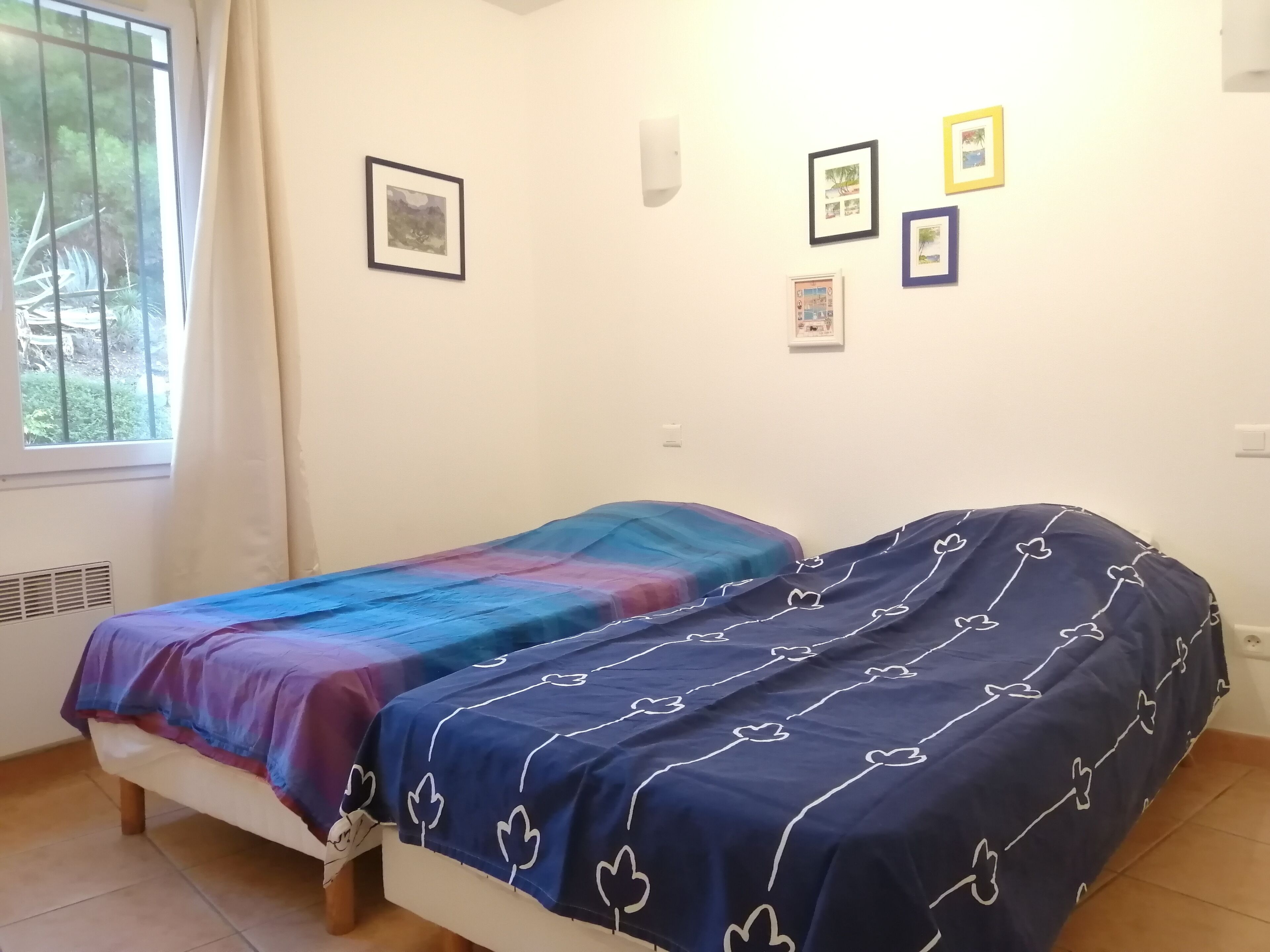 A bedroom with two beds, a window with curtains, and several framed pictures on the wall.