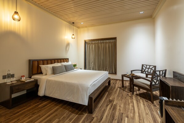 Una habitación de hotel moderna con piso de madera, una cama grande con sábanas blancas, una mesita de noche de madera, un escritorio con una silla y un ventanal con cortinas.