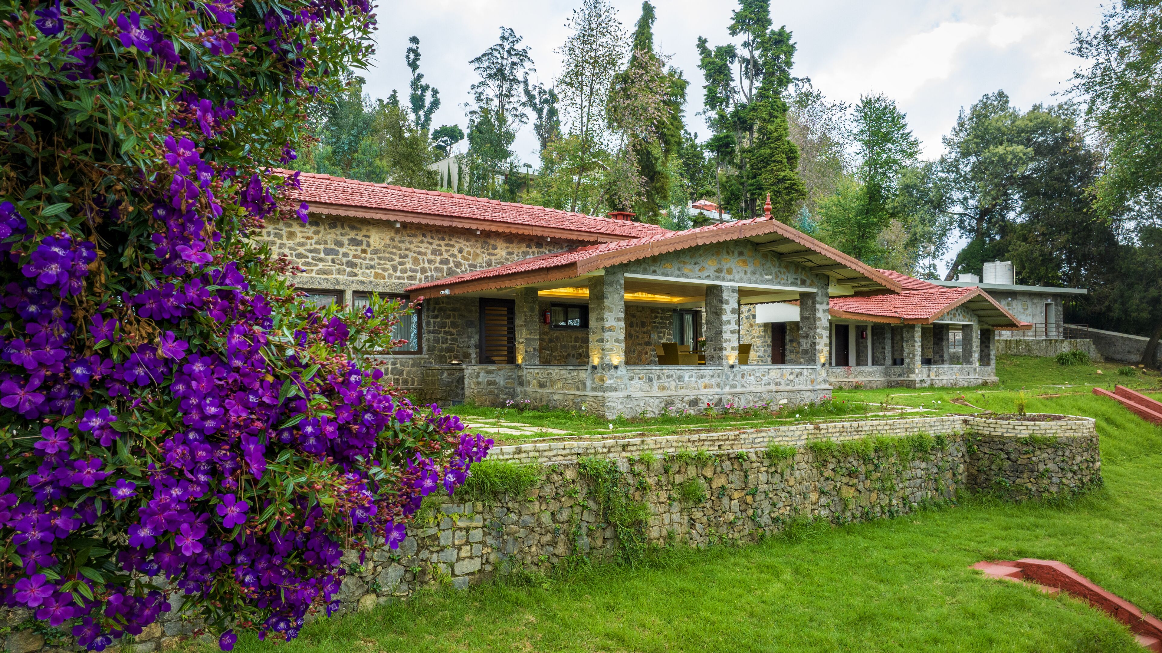 A stone house with a red-tiled roof, surrounded by lush greenery and purple flowers.