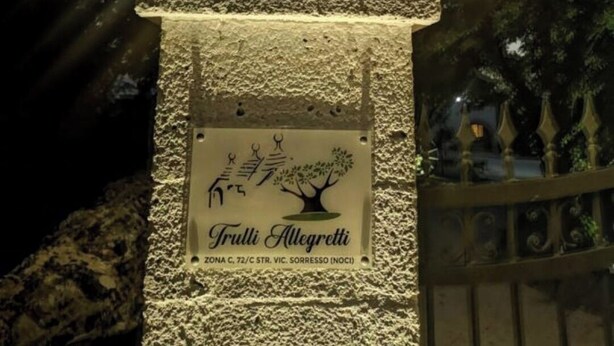 Un monument en pierre, surmonté d’une plaque et d’une structure métallique décorative en forme de couronne, le tout illuminé par des jeux de lumière.
